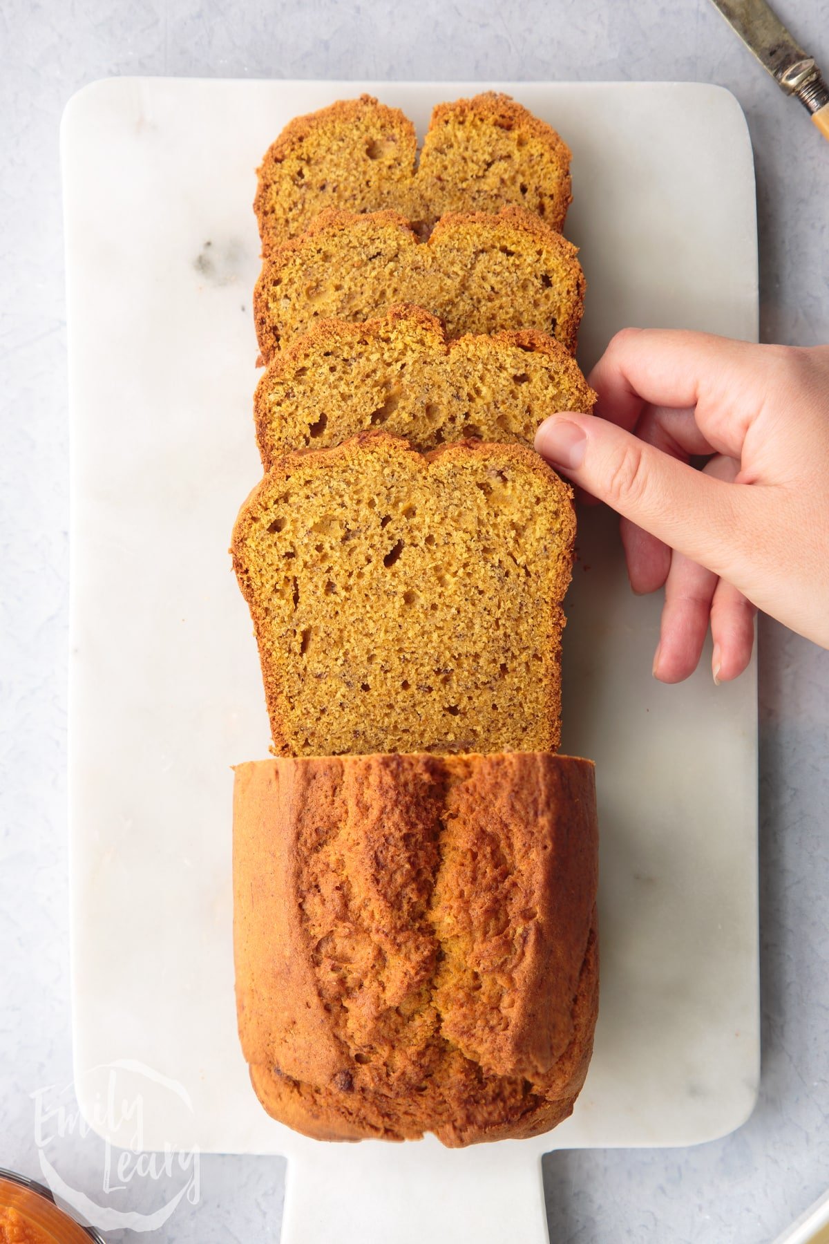 Pumpkin banana bread loaf on a white marble board. Some has been sliced. A hand reaches to take one.