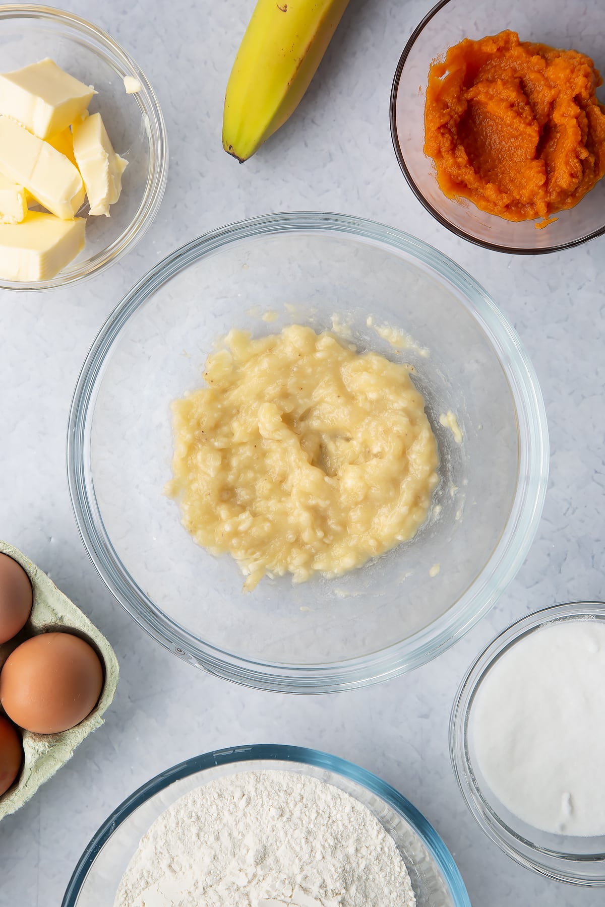 Mashed banana in a glass mixing bowl. Ingredients to make pumpkin banana bread surround the bowl.