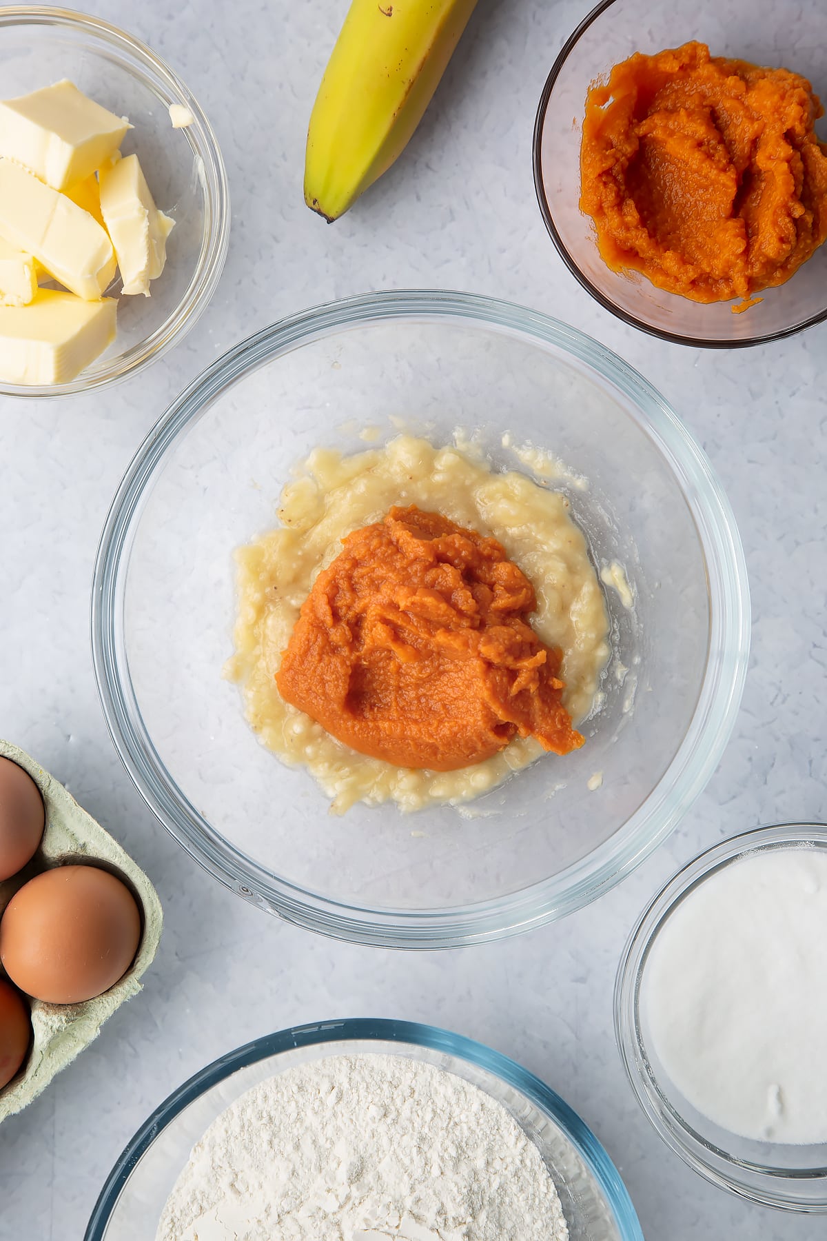 Mashed banana and pumpkin puree in a glass mixing bowl. Ingredients to make pumpkin banana bread surround the bowl.