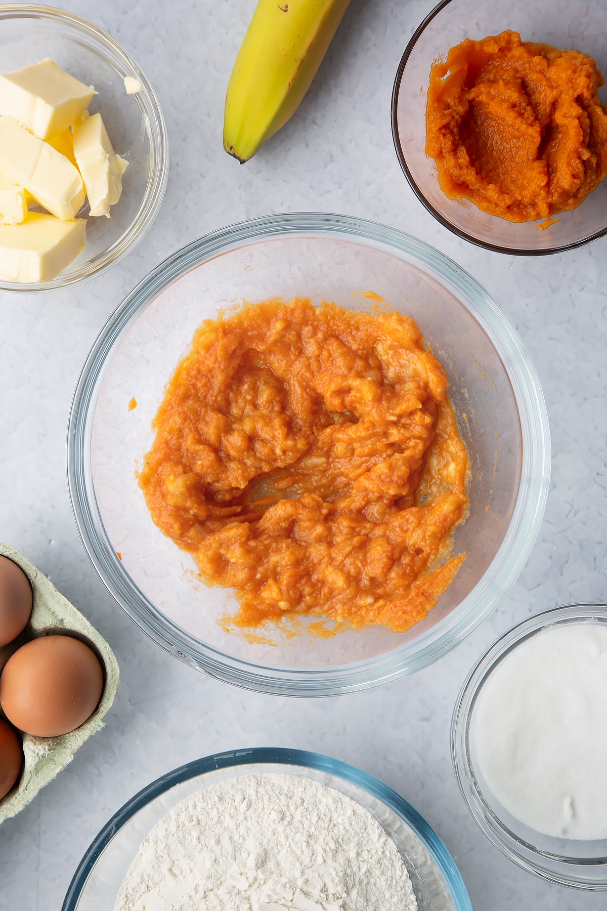 Mashed banana mixed with pumpkin puree in a glass mixing bowl. Ingredients to make pumpkin banana bread surround the bowl.