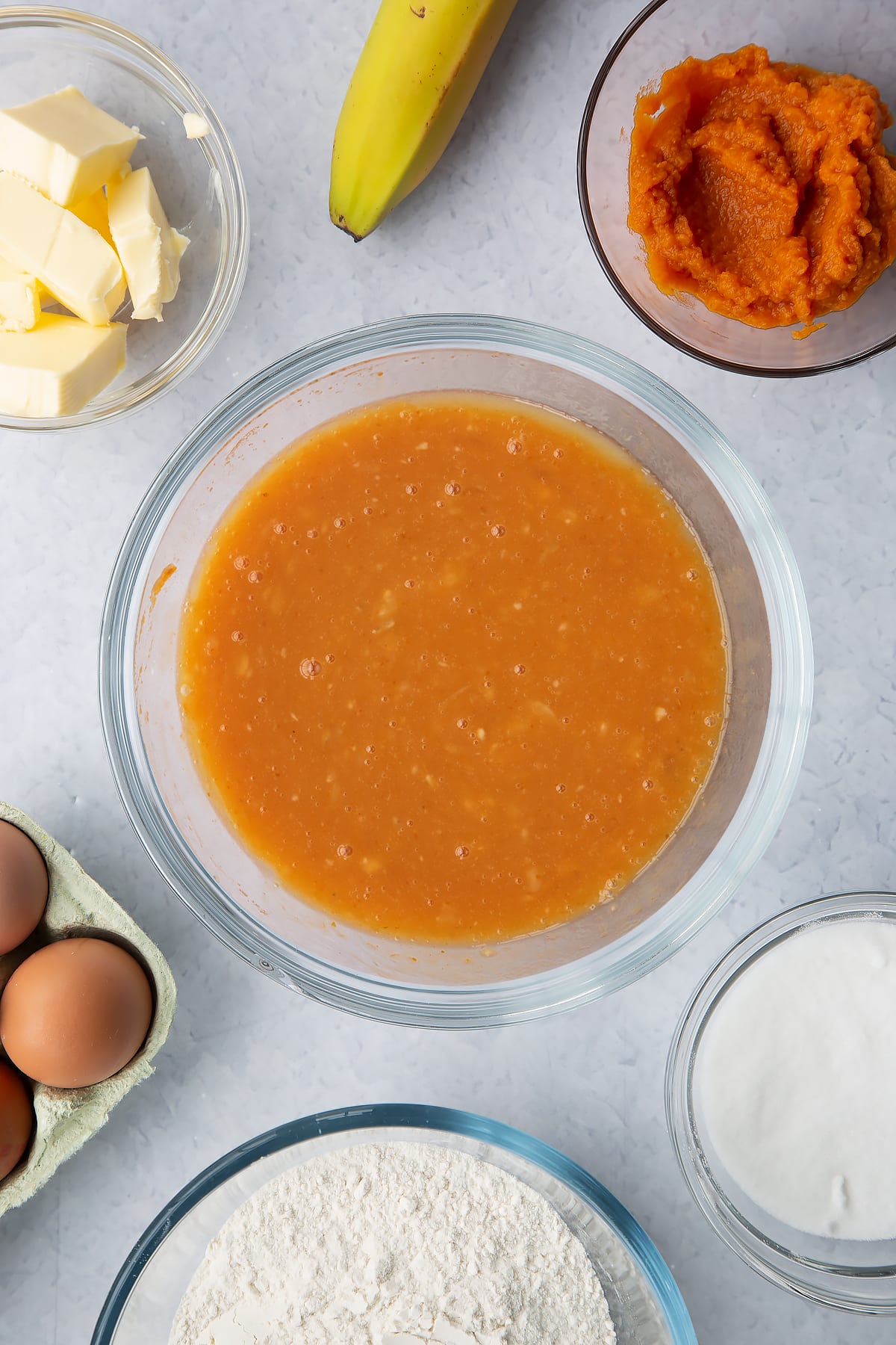 Mashed banana mixed with pumpkin puree, eggs, sugar and melted butter in a glass mixing bowl. Ingredients to make pumpkin banana bread surround the bowl.