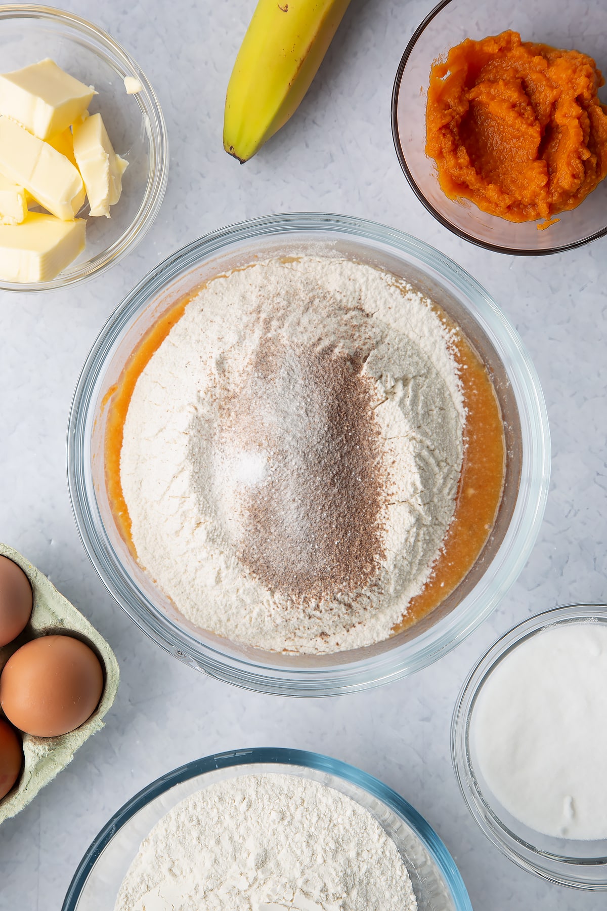 Mashed banana mixed with pumpkin puree, eggs, sugar and melted butter in a glass mixing bowl with flour, baking powder and mixed spice on top. Ingredients to make pumpkin banana bread surround the bowl.