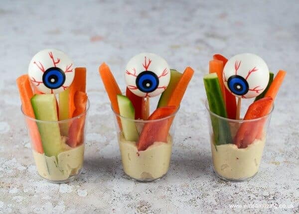 Hummus at the bottom of three plastic cups, topped with fingers of raw carrot, pepper and cucumber. Decorative plastic eyeballs have been added to each cup.