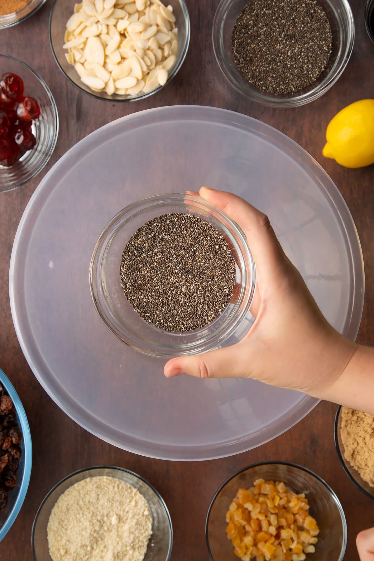 Hand holding a small bowl with chia seeds. Beneath is a larger bowl, surrounded by ingredients to make vegan Christmas cake.