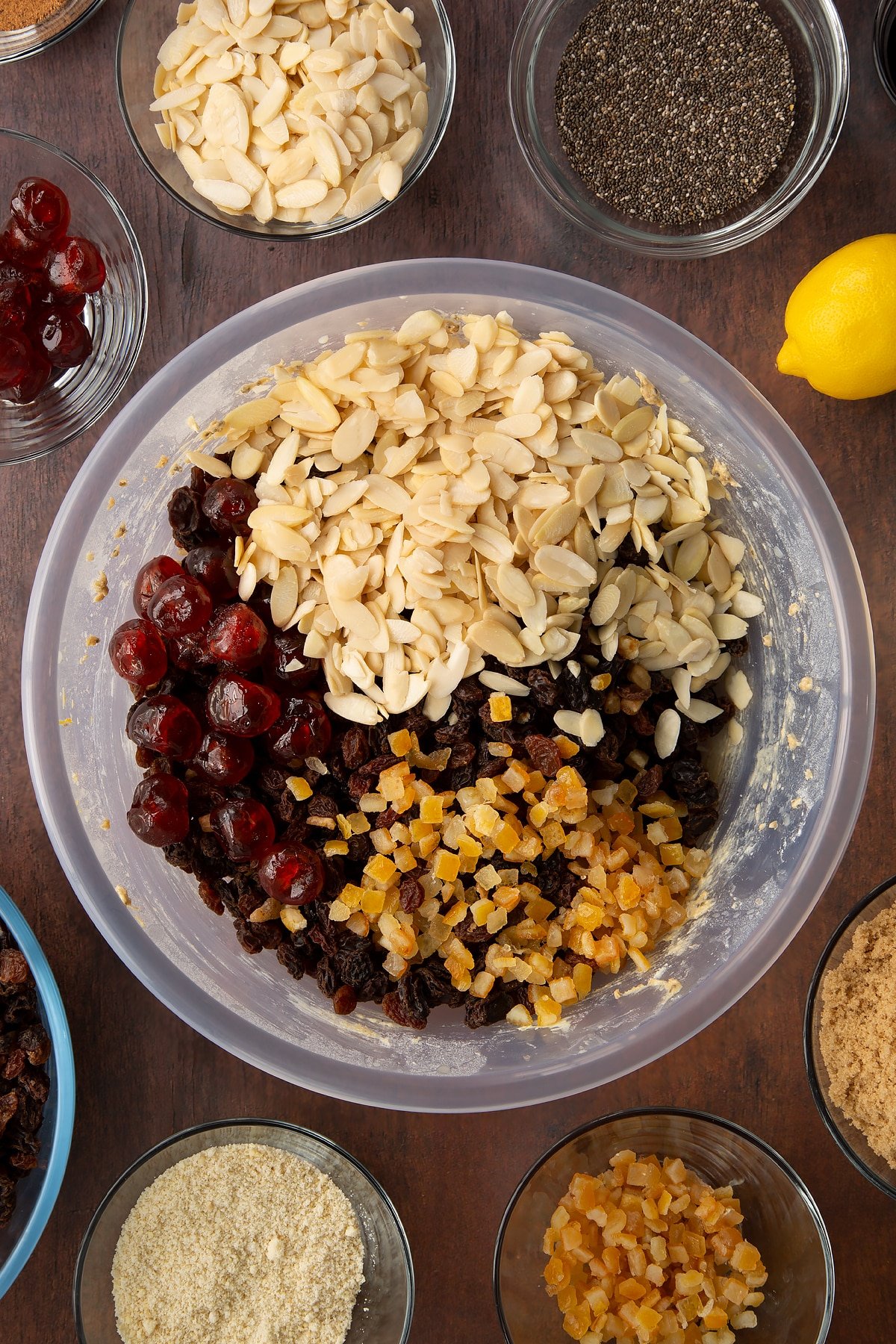 Butter, sugar, treacle, flour, almonds, spices, citrus juice and plant milk in a bowl with flaked almonds, cherries and mixed peel on top. Ingredients to make vegan Christmas cake surround the bowl.