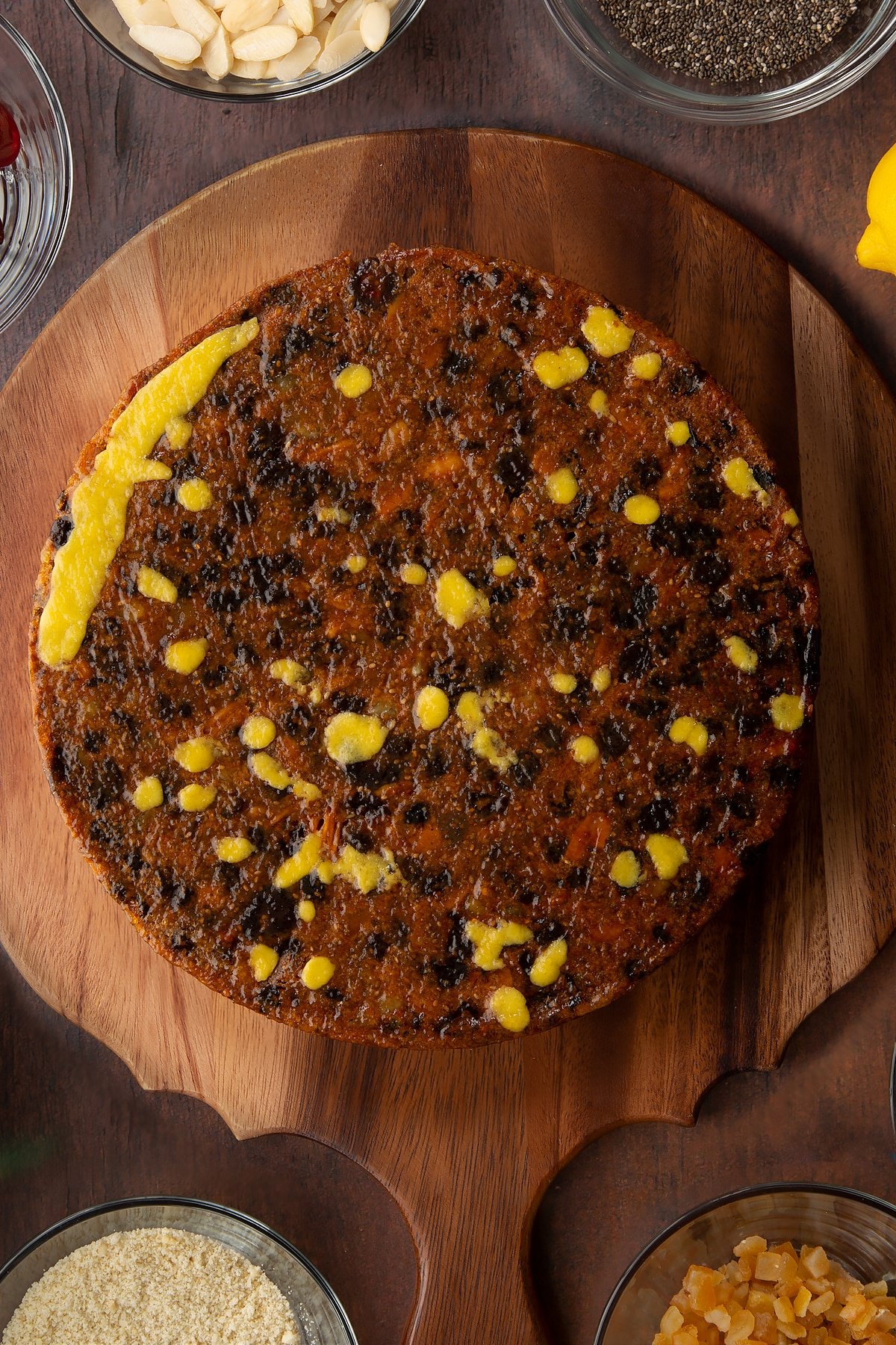 Vegan Christmas cake, upside down on a wooden board. Any dips are filled with small pieces of marzipan. The cake is brushed with apricot jam.