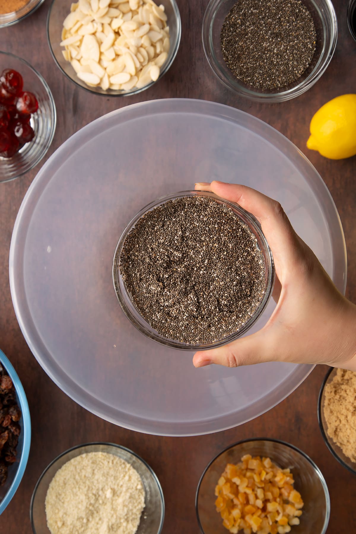 Hand holding a small bowl with chia seeds and water. Beneath is a larger bowl, surrounded by ingredients to make vegan Christmas cake.