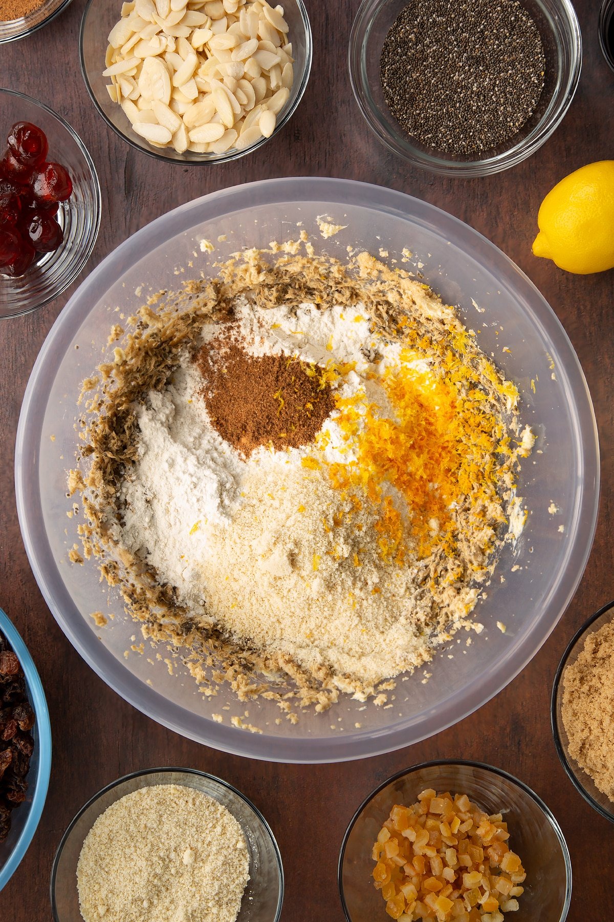 Vegan butter, brown sugar, chia "egg" and black treacle, whisked together in a large plastic bowl with Add the flour, ground almonds, mixed spice, nutmeg, orange zest and lemon zest on top. Ingredients to make vegan Christmas cake surround the bowl.