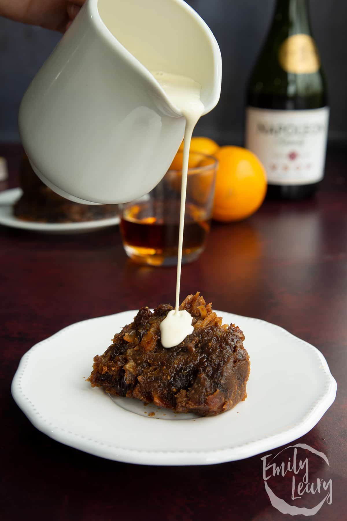 A piece of vegan Christmas pudding on a small white plate. Vegan cream is being poured on to it.