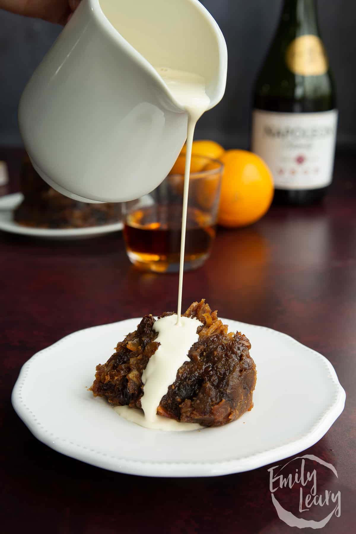 A piece of vegan Christmas pudding on a small white plate. Vegan cream is being poured on to it and running onto the plate