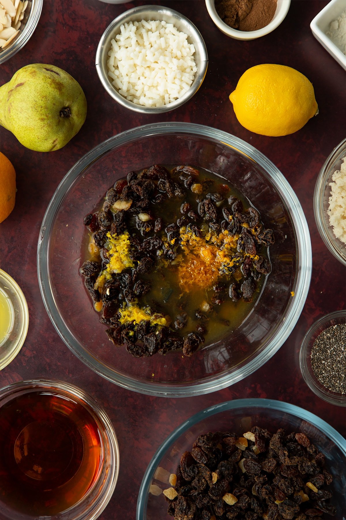 Mixed fruit, brandy, citrus juice and zest in a glass mixing bowl. Ingredients to make vegan Christmas pudding surround the bowl.