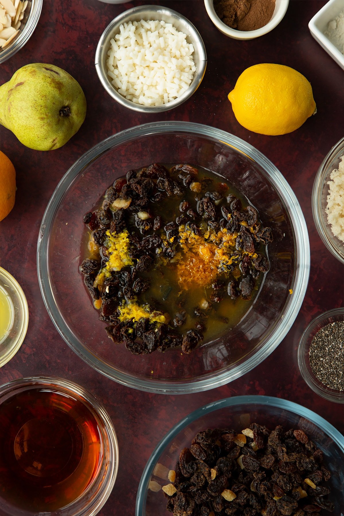 Mixed fruit, brandy, citrus juice and zest in a glass mixing bowl. Ingredients to make vegan Christmas pudding surround the bowl. 