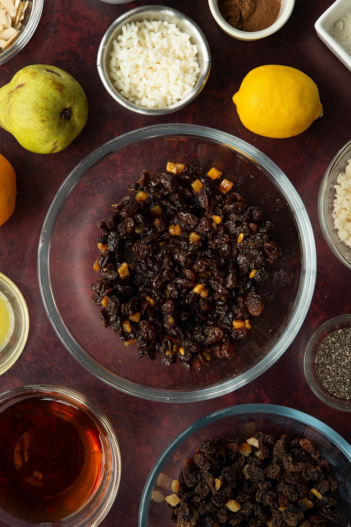 Mixed fruit cooked in brandy, citrus juice and zest in a glass mixing bowl. Ingredients to make vegan Christmas pudding surround the bowl.