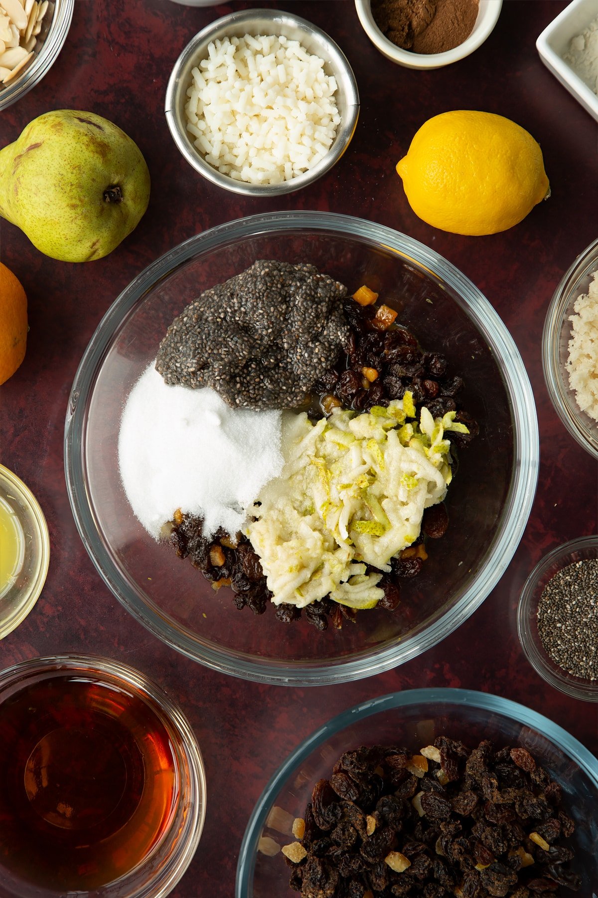 Mixed fruit cooked in brandy, citrus juice and zest in a glass mixing bowl with grated pear, soaked chia seeds, black treacle and sugar on top. Ingredients to make vegan Christmas pudding surround the bowl.