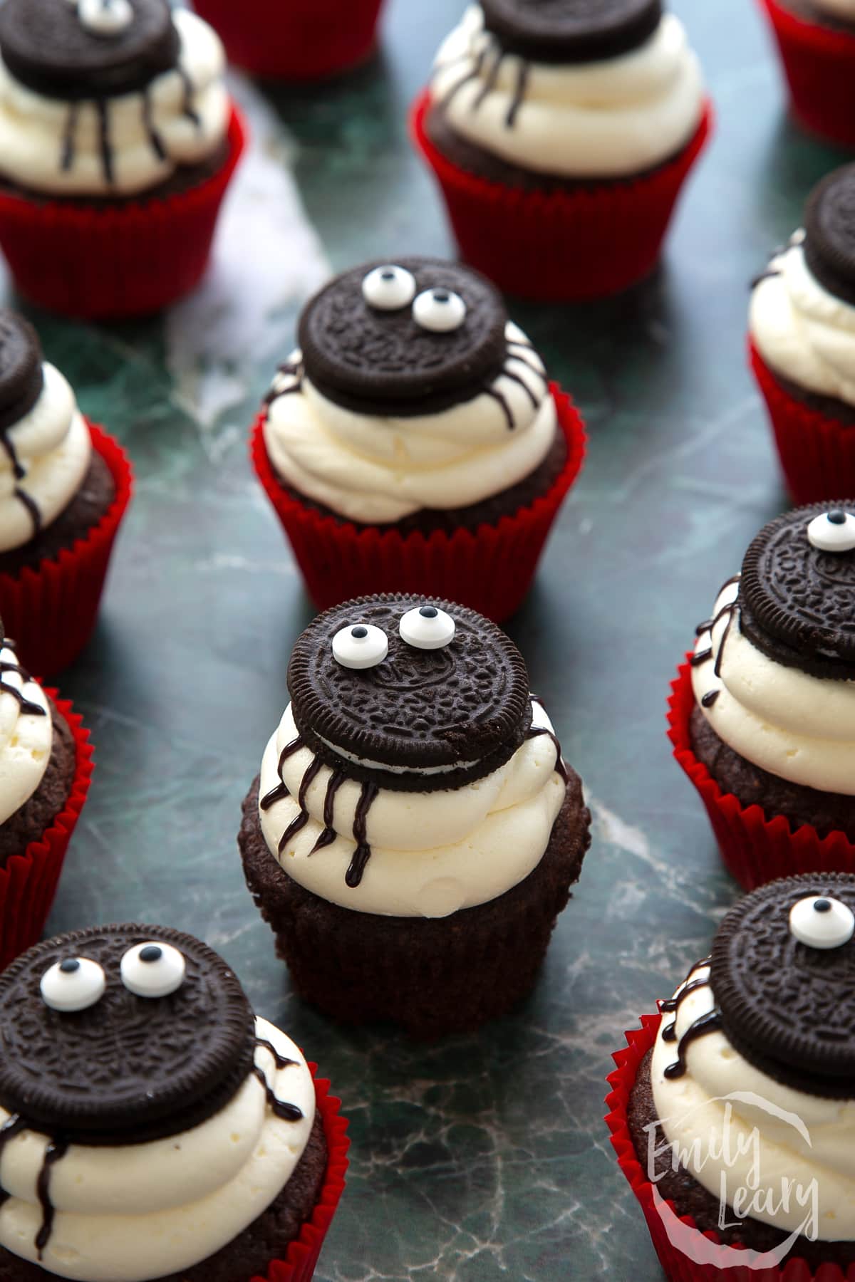 A vegan Halloween cupcake, decorated with Oreos and candy eyes to look like a spider. It is unwrapped and surrounded by more cupcakes.