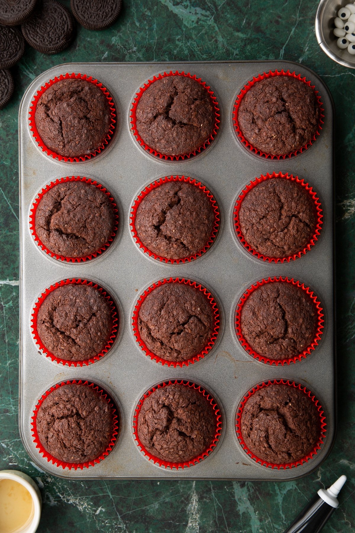Freshly baked vegan chocolate cupcakes in a 12-hole muffin tray. Ingredients to make vegan Halloween cupcakes surround the tray.