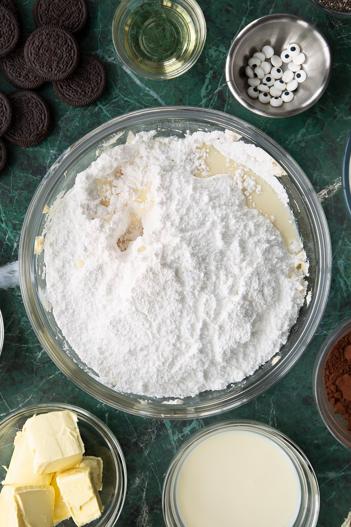 Whipped vegan butter in a glass mixing bowl topped with icing sugar, vanilla and plant milk. Ingredients to make vegan Halloween cupcakes surround the bowl.