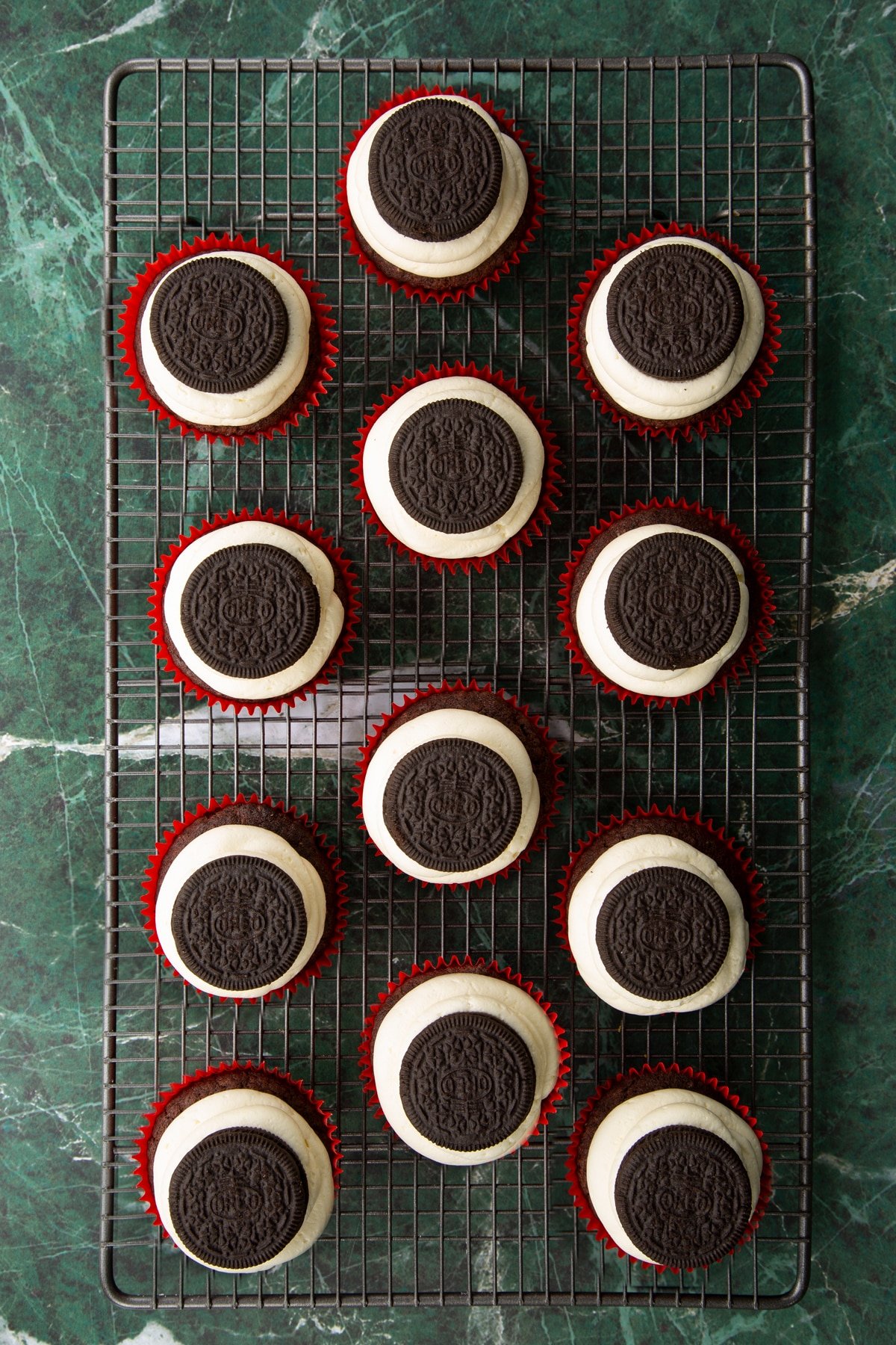 Vegan chocolate cupcakes on a wire cooling rack. They have vanilla frosting piped on top, with an Oreo on top of each one.