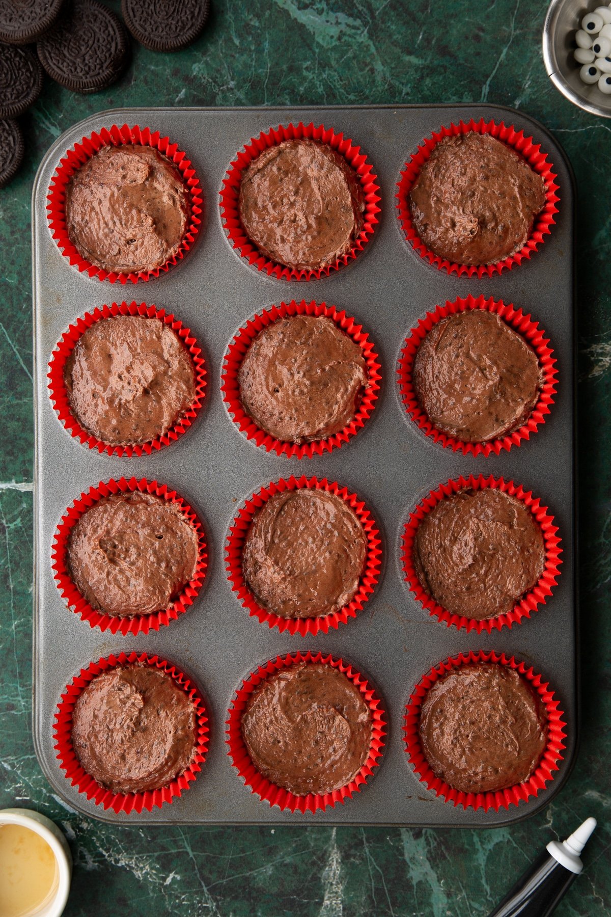 Cupcake cases filled with vegan chocolate cake batter in a 12-hole muffin tray. Ingredients to make vegan Halloween cupcakes surround the tray.