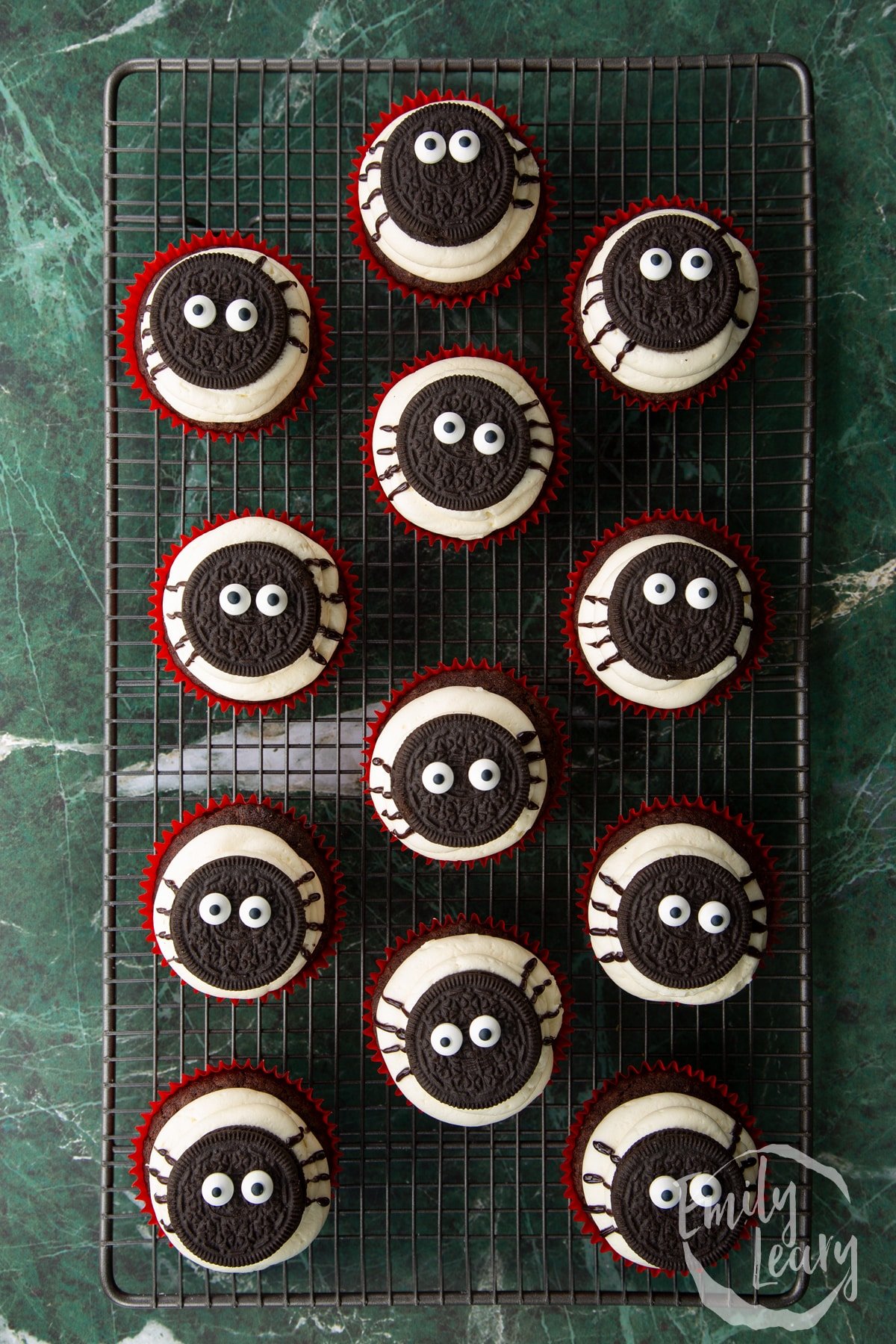 Vegan chocolate cupcakes on a wire cooling rack. They have vanilla frosting piped on top, then an Oreo with candy eyes. Spider legs are piped on with a black icing pen.