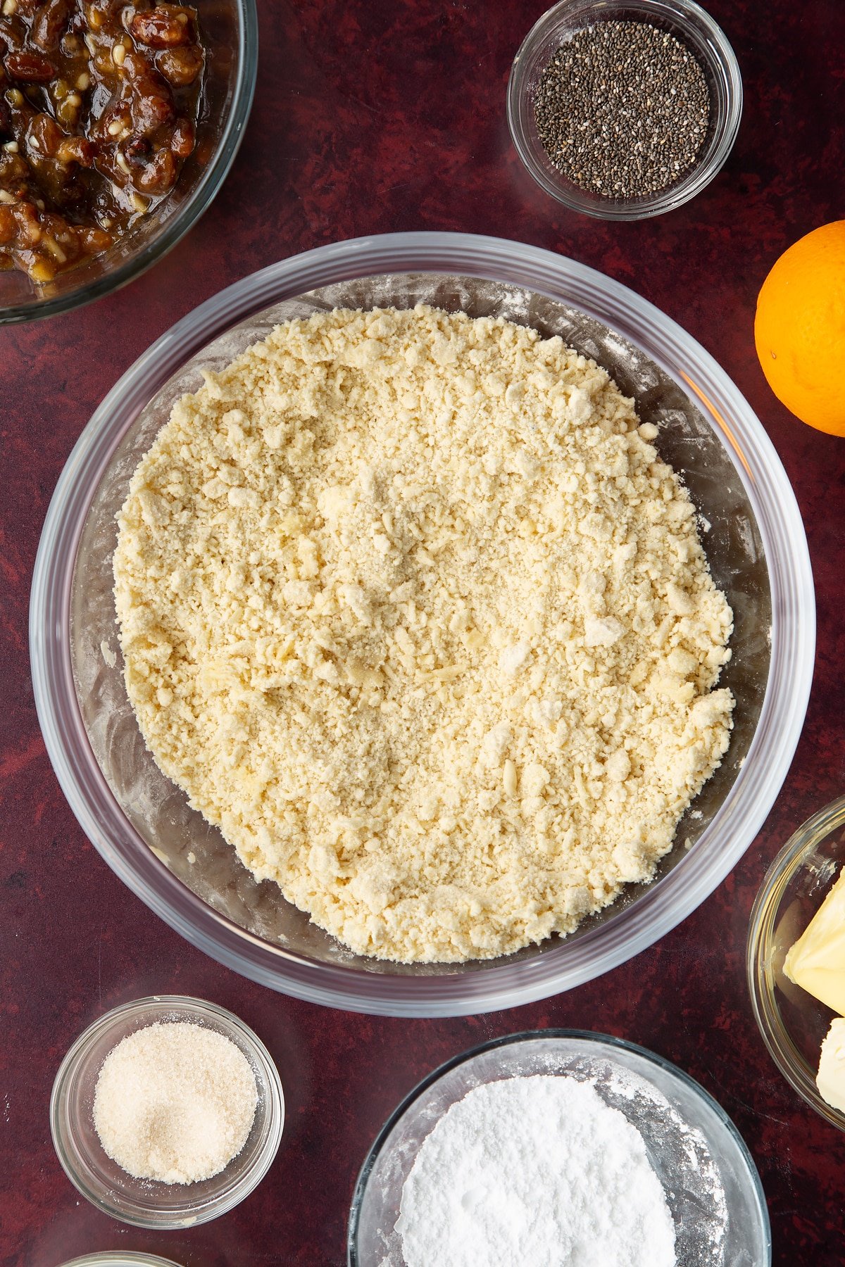 Flour, icing sugar and vegan butter rubbed together in a bowl. Ingredients to make a vegan mince pie recipe surround the bowl.