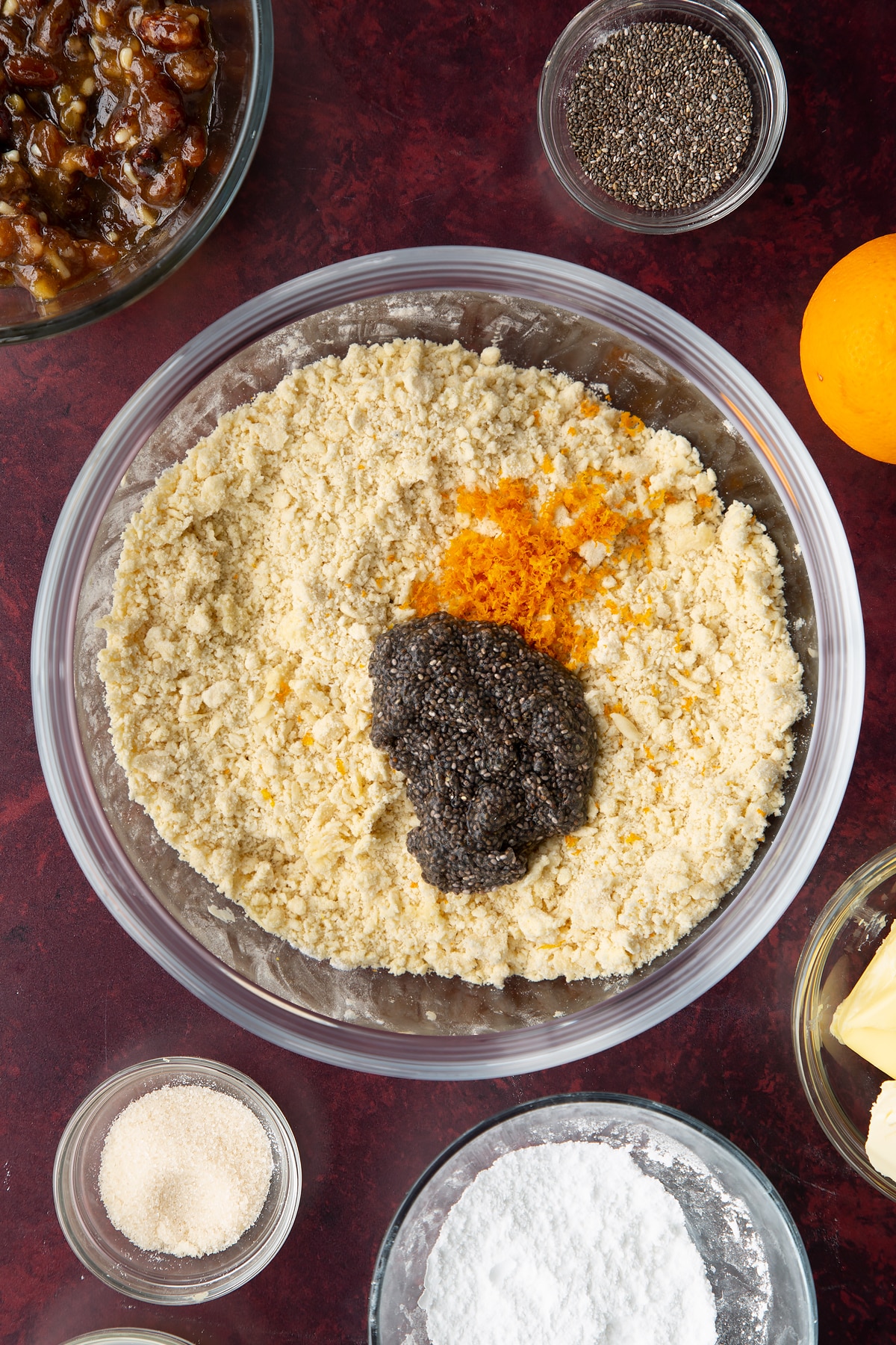 Flour, icing sugar and vegan butter rubbed together in a bowl with chia egg and orange zest on top. Ingredients to make a vegan mince pie recipe surround the bowl.