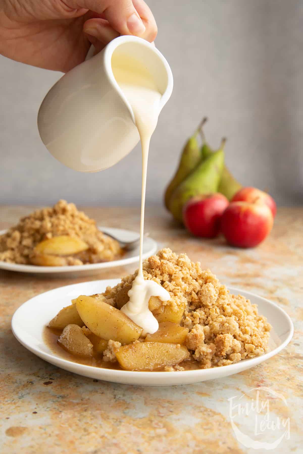 Apple pear crumble served to a small white plate. A hand pours a jug of custard above the plate.