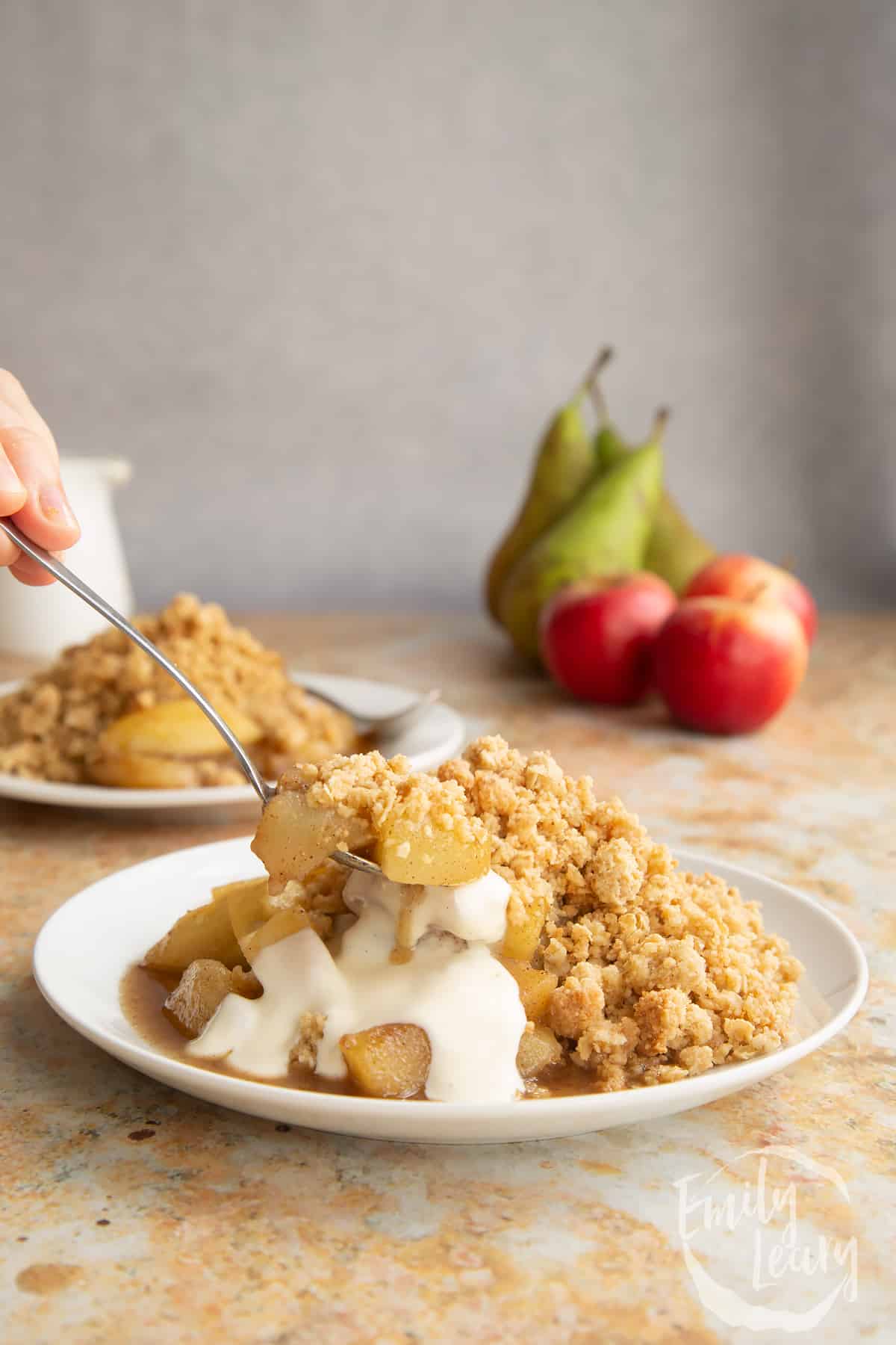 Apple pear crumble served to a small white plate with some custard. A small spoon delves in.