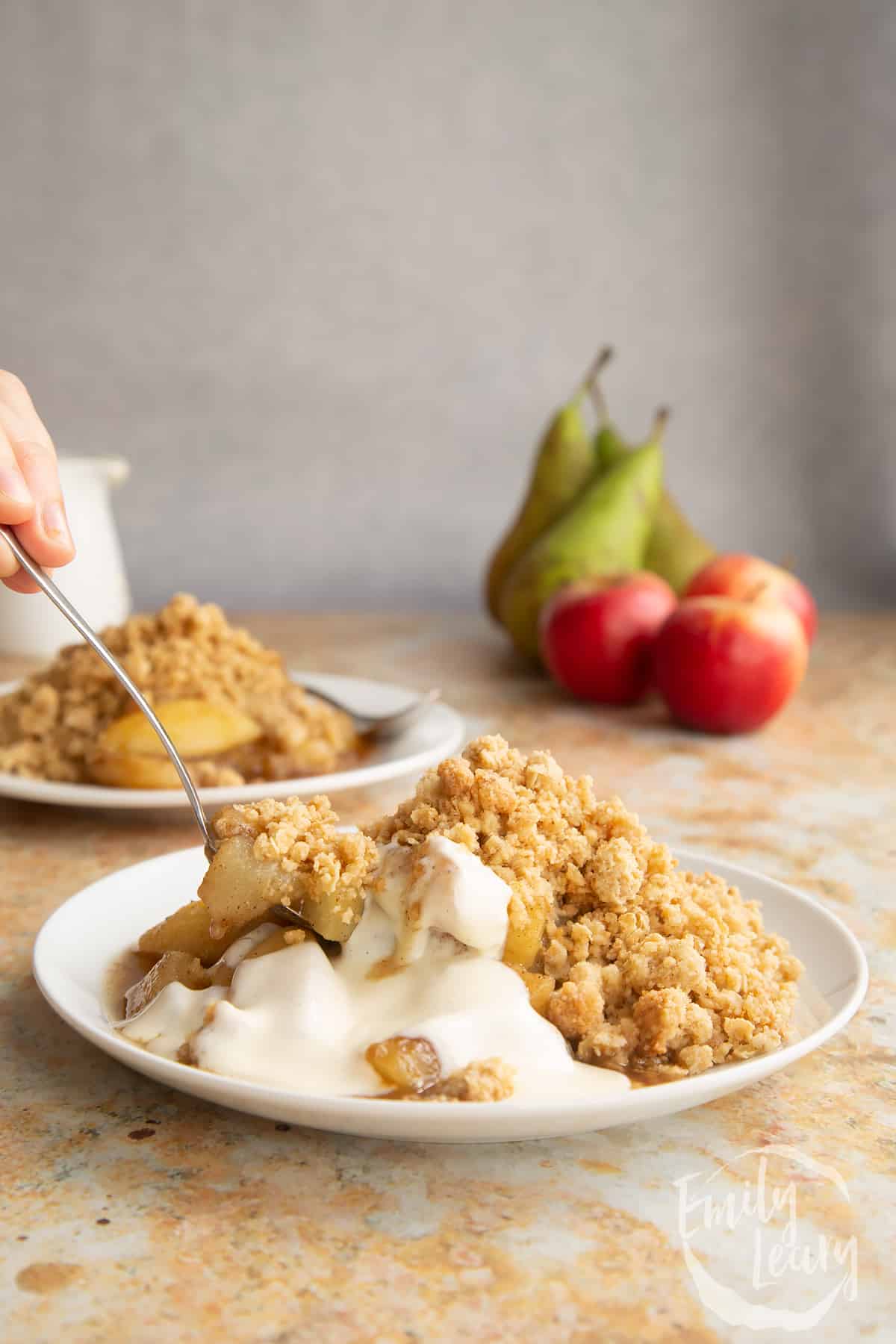 Apple pear crumble and custard served to a small white plate. A hand holds a spoon delving into the crumble.