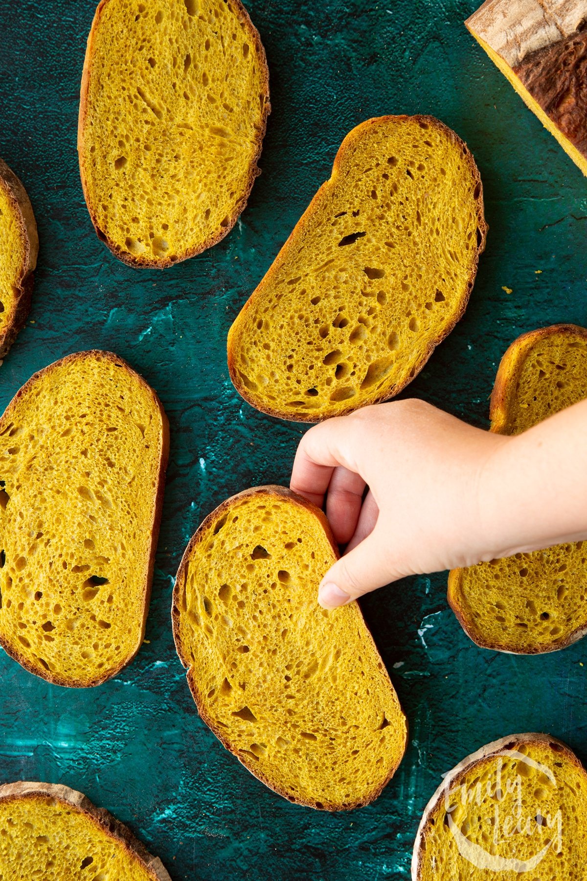 Slices of pumpkin sourdough bread arranged on a dark green surface. A hand reaches for one.