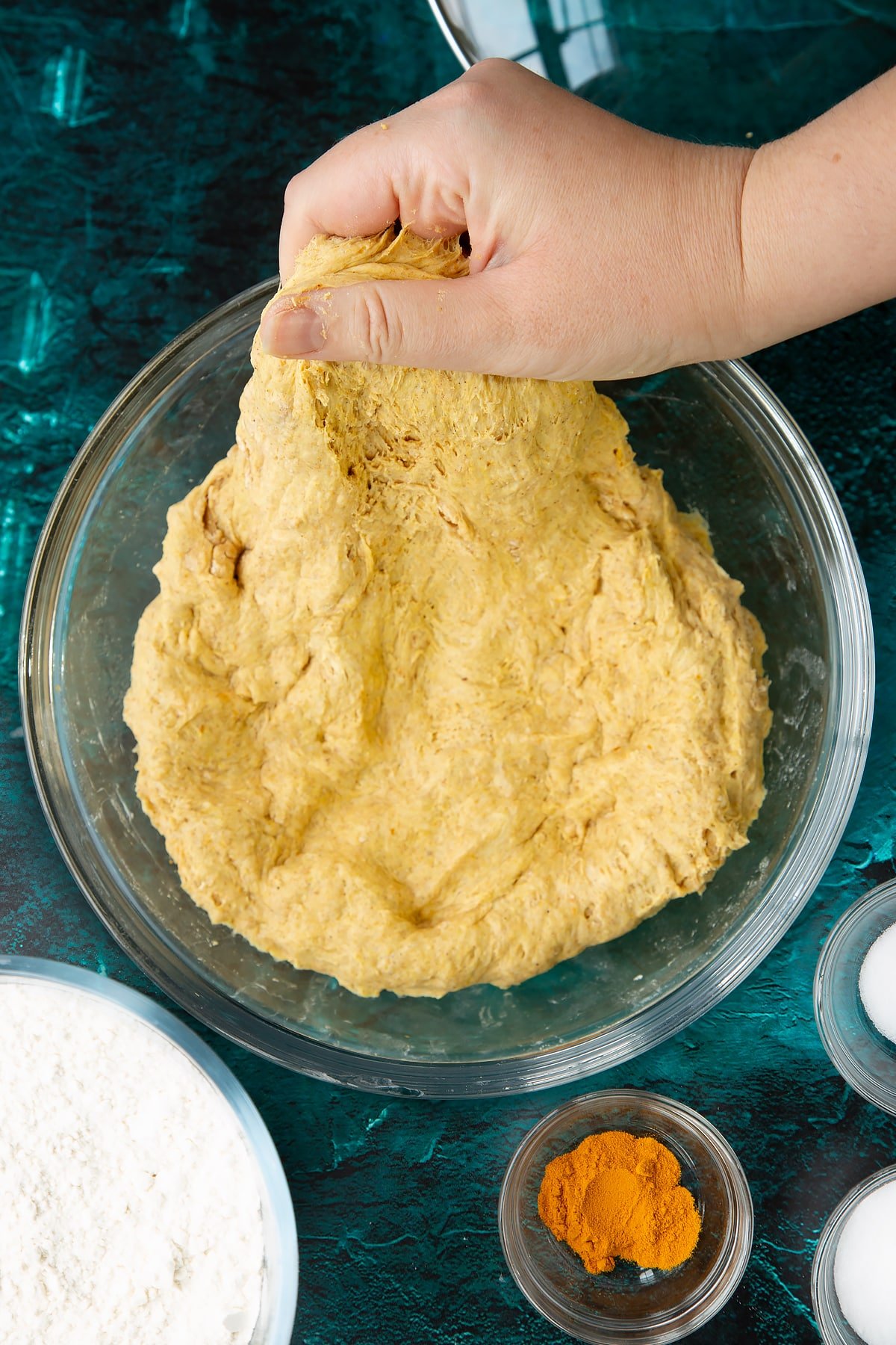 Pumpkin sourdough pressed into the base of a bowl. A hand stretches some out. Ingredients to make pumpkin sourdough bread surround the bowl.