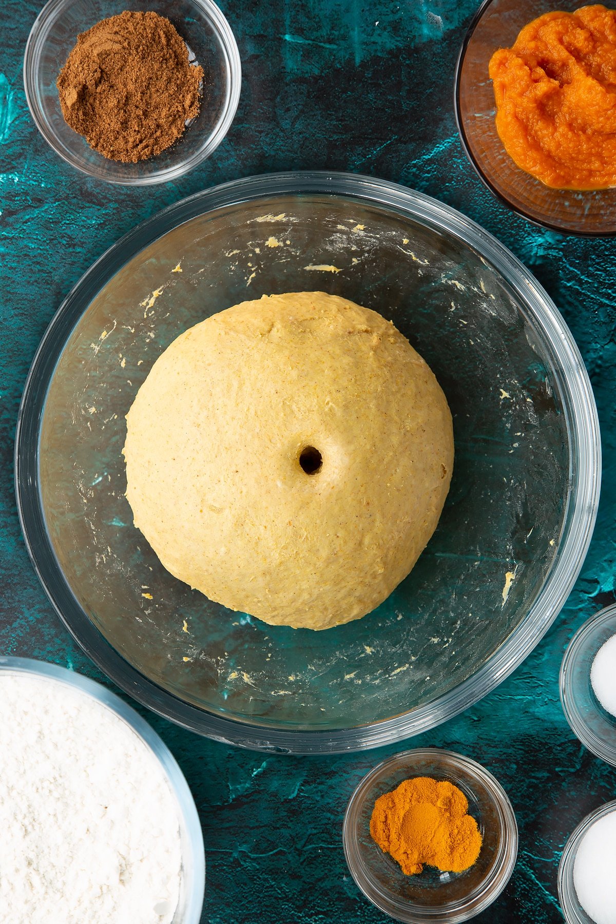 A ball of pumpkin sourdough with one hole on top in a bowl. Ingredients to make pumpkin sourdough bread surround the bowl.