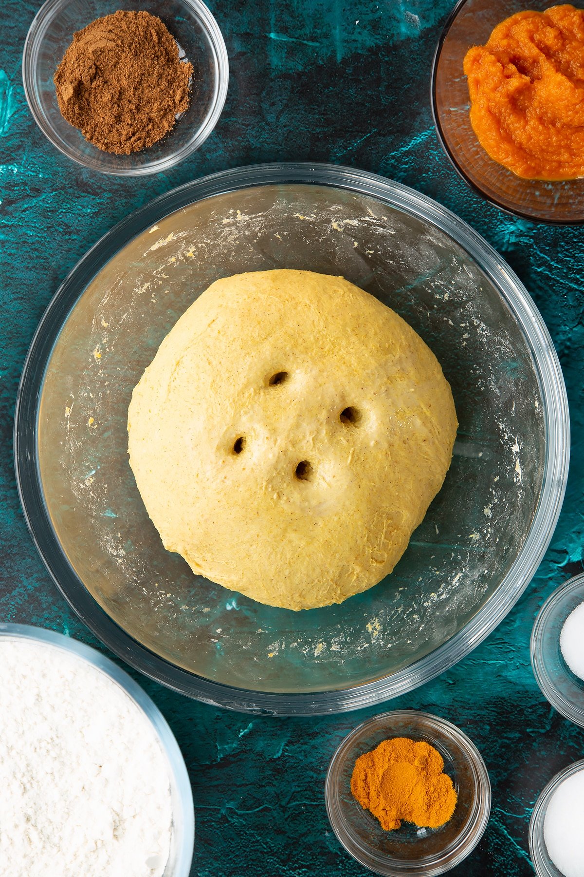 A ball of pumpkin sourdough with four holes on top in a bowl. Ingredients to make pumpkin sourdough bread surround the bowl.