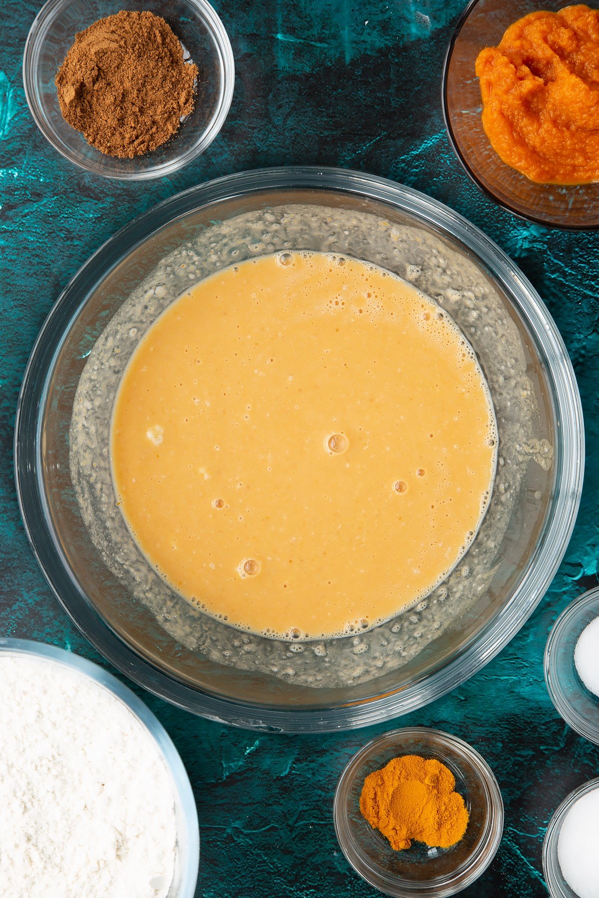 Pumpkin puree, warm water and sourdough preferment, whisked together in a bowl. Ingredients to make pumpkin sourdough bread surround the bowl.