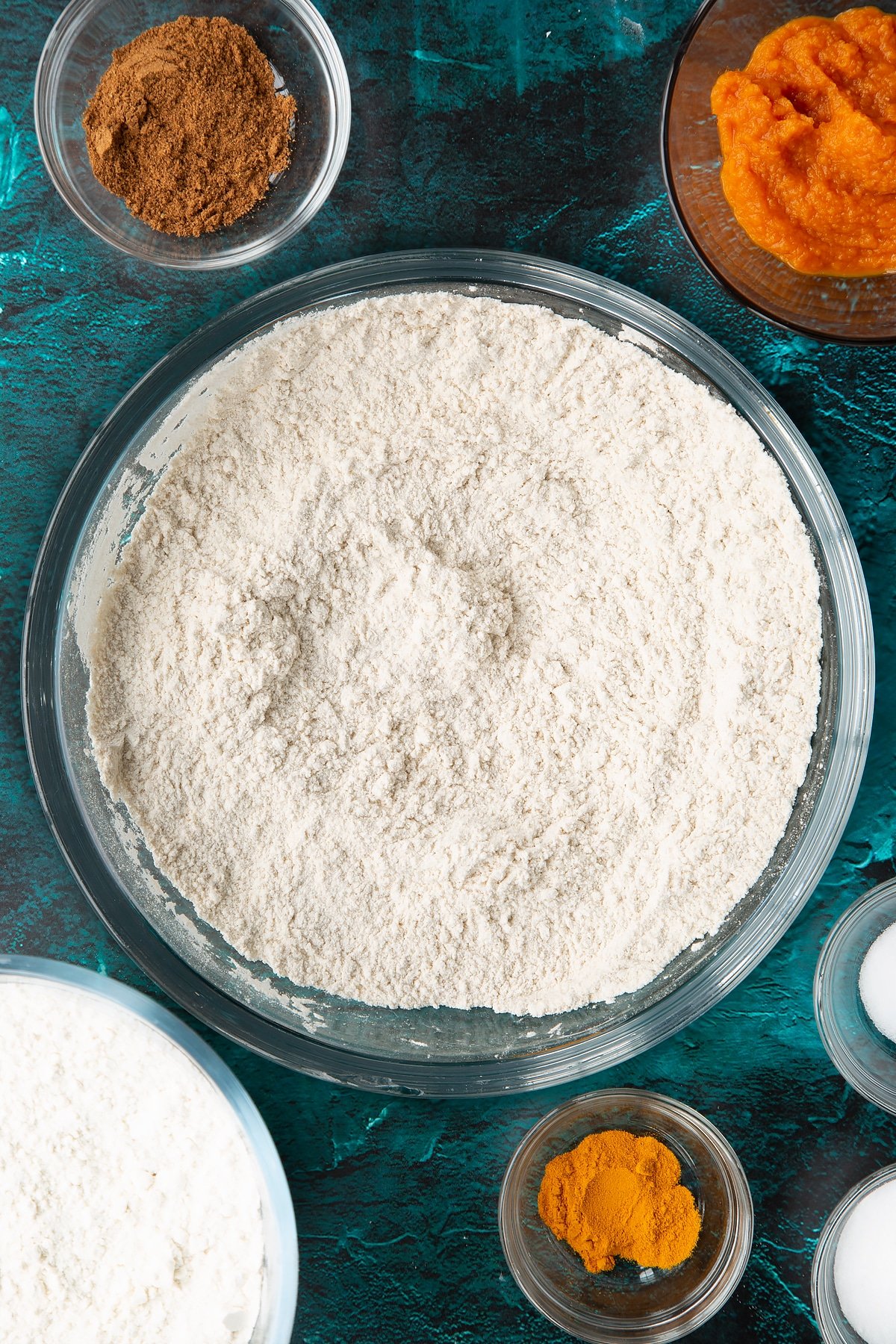 Flour, sugar, salt, mixed spice and turmeric, whisked together in a bowl. Ingredients to make pumpkin sourdough bread surround the bowl.