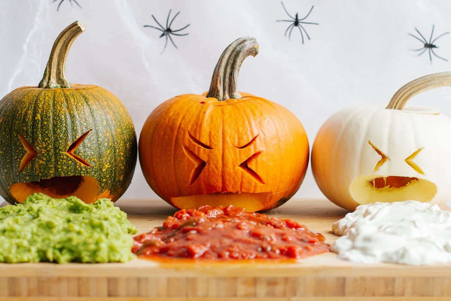 A row of three pumpkins. Each have cartoonish facial expressions and appear to be vomiting different taco sauces onto a board.