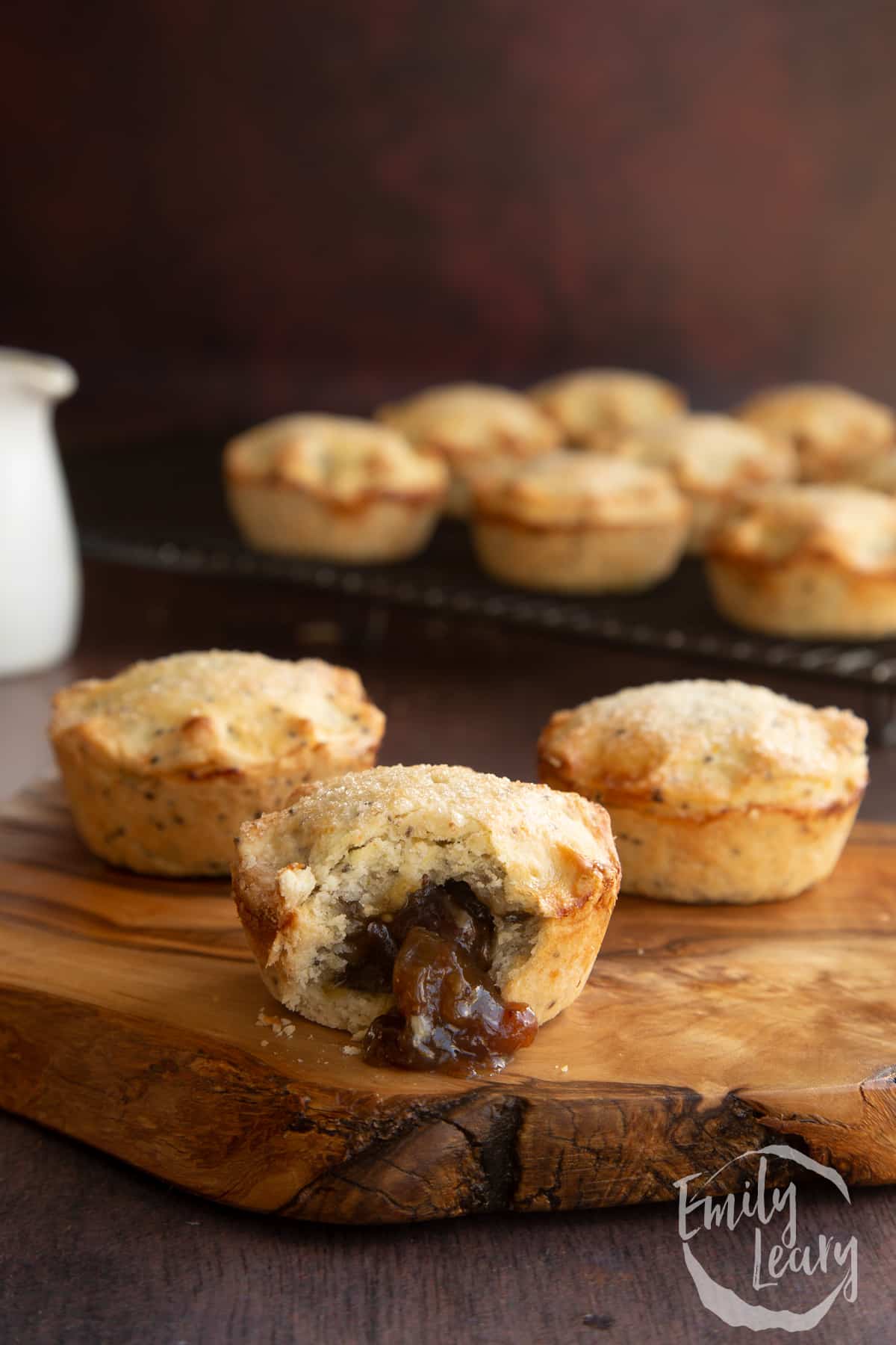 Vegan mince pies on a olive board with more on a wire rack in the background. One has been bitten open.