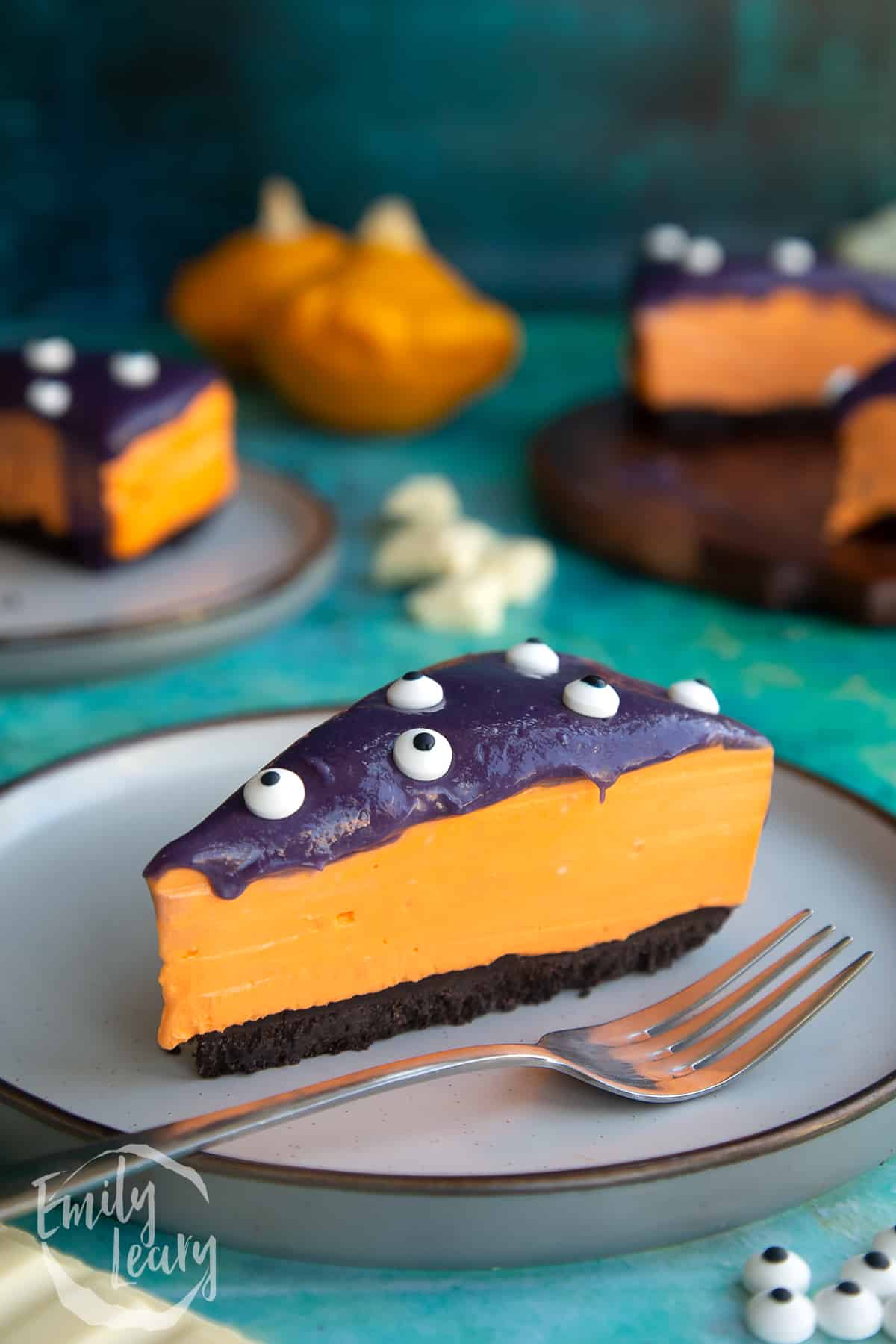 Slice of orange and strawberry Halloween cheesecake with an Oreo base on a plate with a fork.