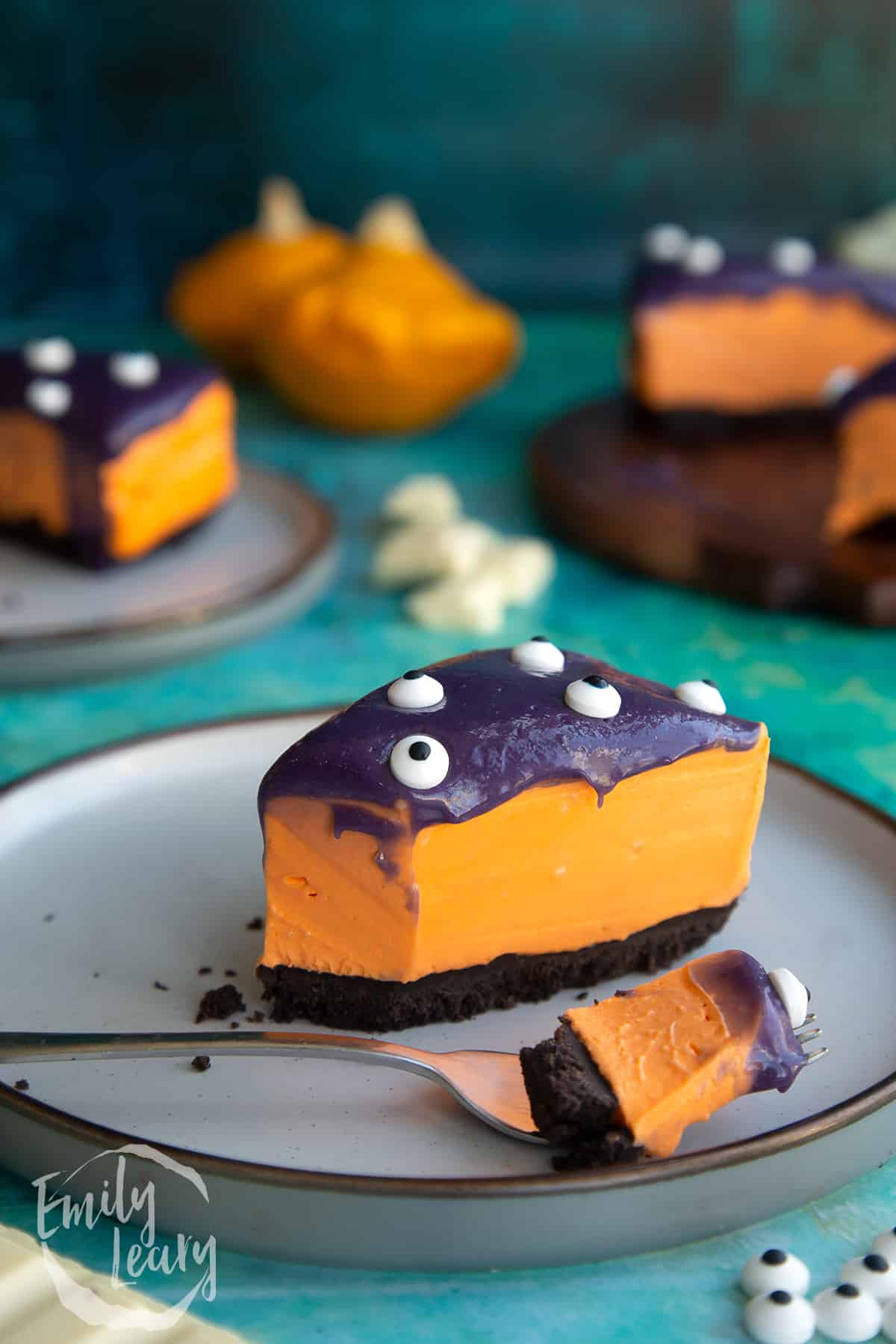 Slice of orange and strawberry Halloween cheesecake with an Oreo base on a plate. Some has been removed with a fork that rests on the plate.