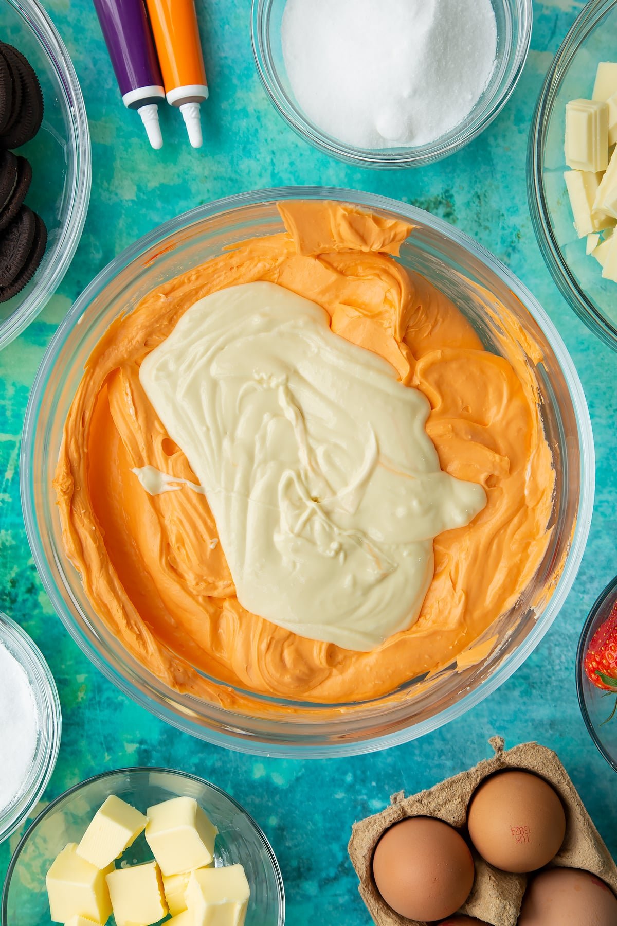 Orange cheesecake mix topped with white chocolate in a bowl. Ingredients to make Halloween cheesecake surround the bowl.