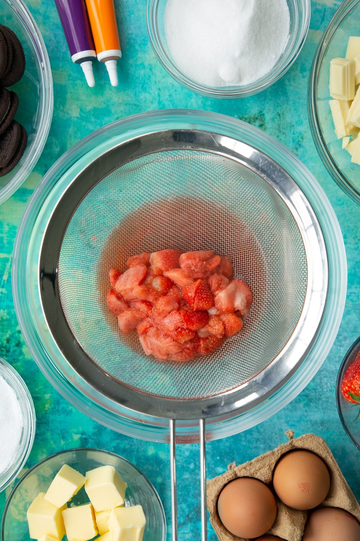 Cooked strawberries in a sieve over a bowl. Ingredients to make Halloween cheesecake surround the bowl.