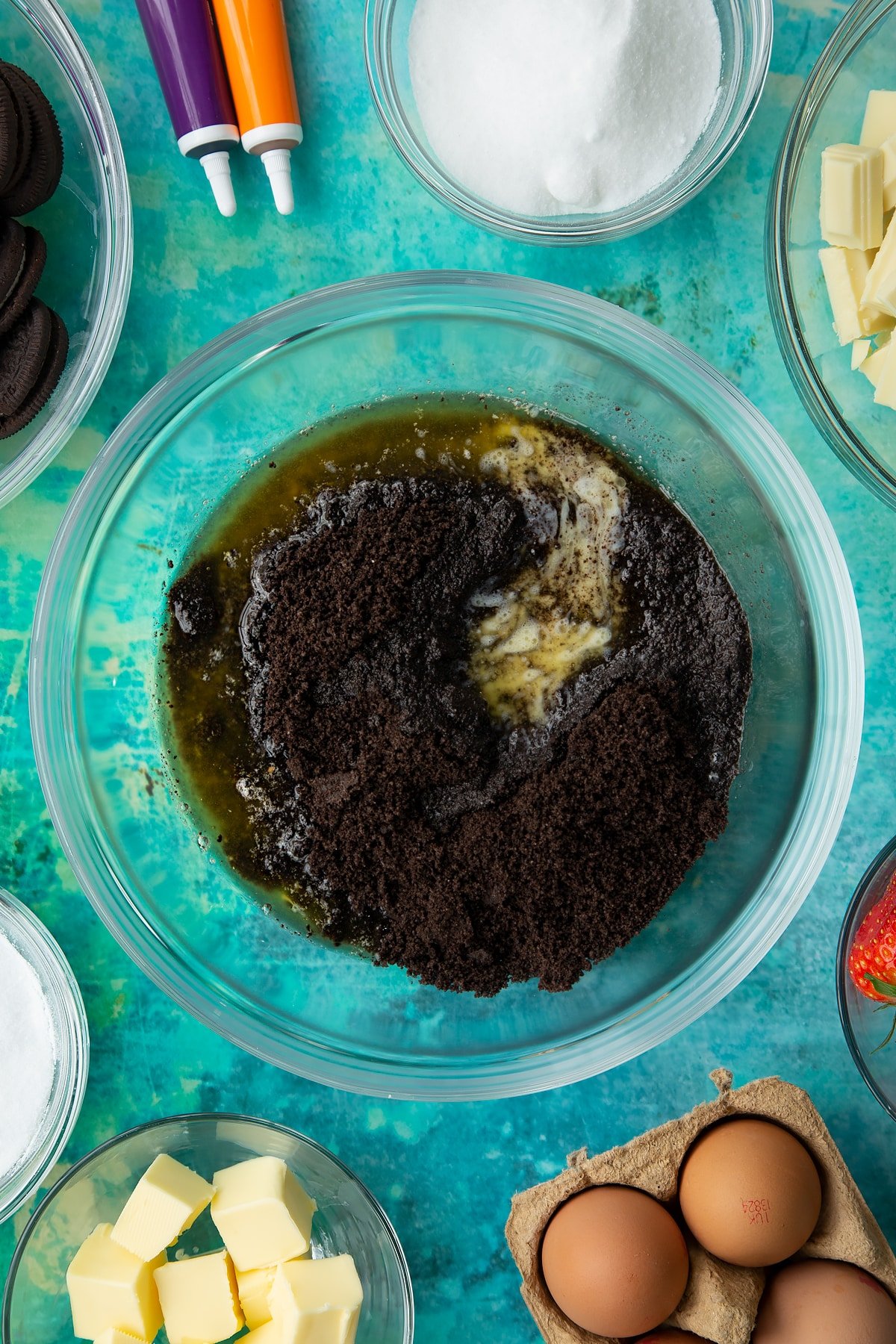 Oreo biscuit crumb and melted butter in a bowl. Ingredients to make Halloween cheesecake surround the bowl.