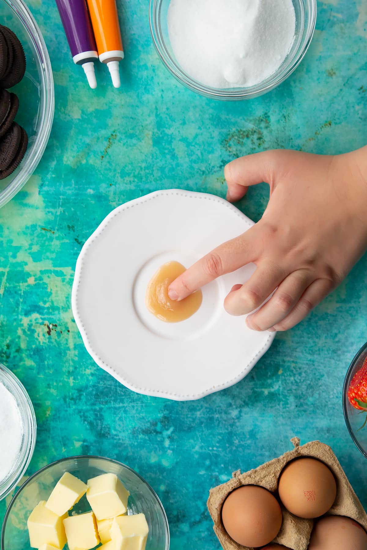 Strawberry curd on a white plate with a finger wrinkling it. Ingredients to make Halloween cheesecake surround the plate.