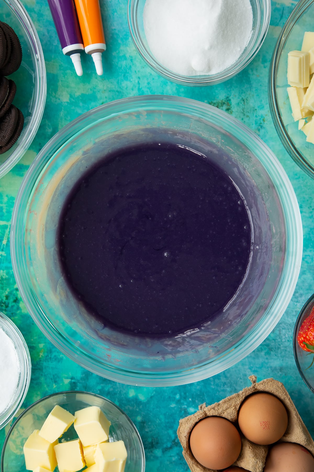 Purple dyed strawberry curd in a bowl. Ingredients to make Halloween cheesecake surround the bowl.