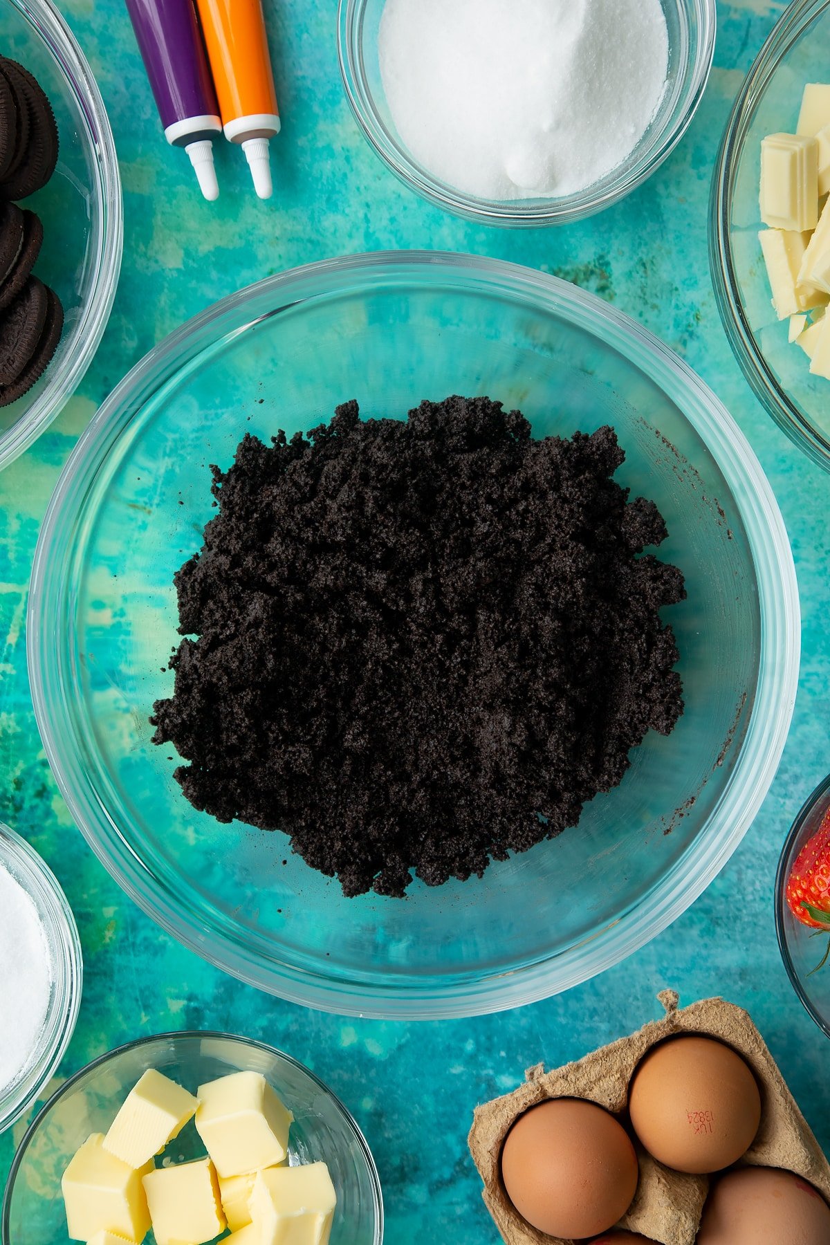 Oreo biscuit crumb mixed with melted butter in a bowl. Ingredients to make Halloween cheesecake surround the bowl.