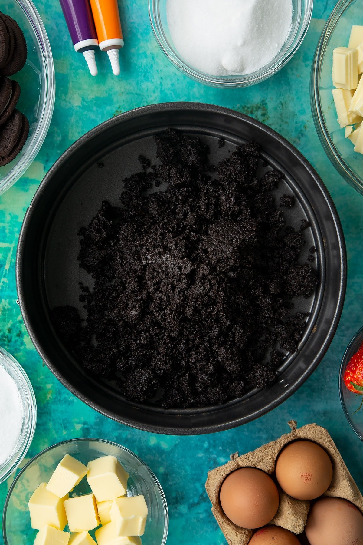 Oreo biscuit crumb mixed with melted butter, tipped into a tin. Ingredients to make Halloween cheesecake surround the tin.