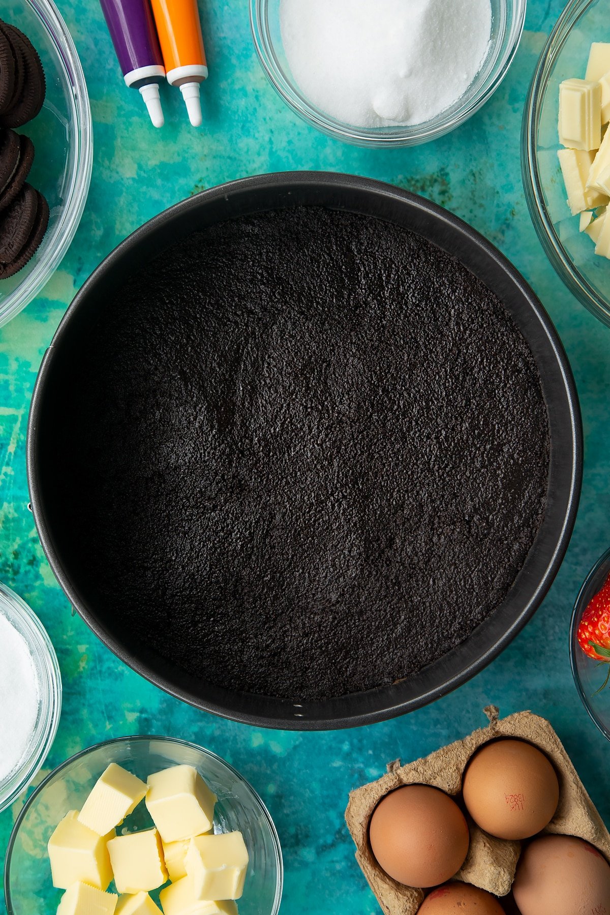 Oreo biscuit pressed into the base of a tin. Ingredients to make Halloween cheesecake surround the tin.