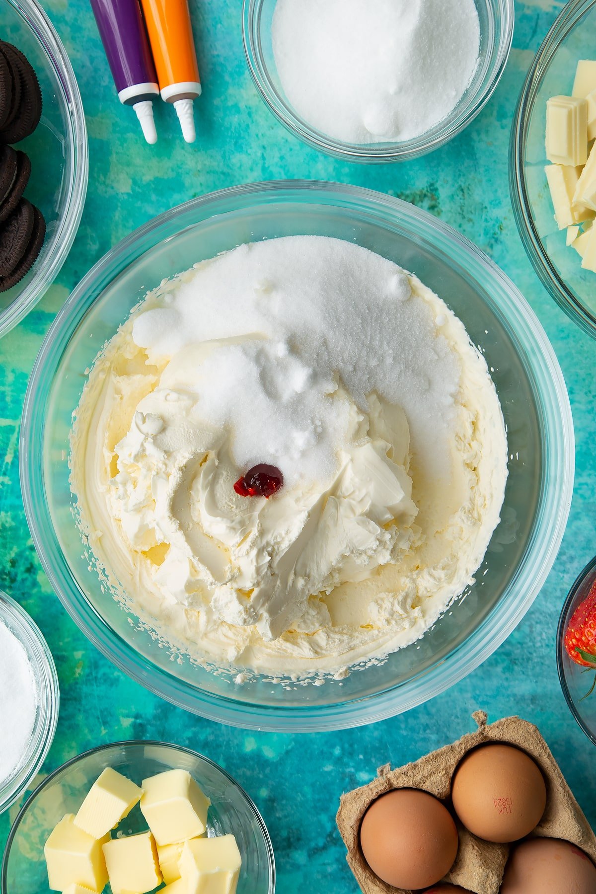 Whipped cream, cream cheese and sugar with orange extract and orange food dye in a bowl. Ingredients to make Halloween cheesecake surround the bowl.