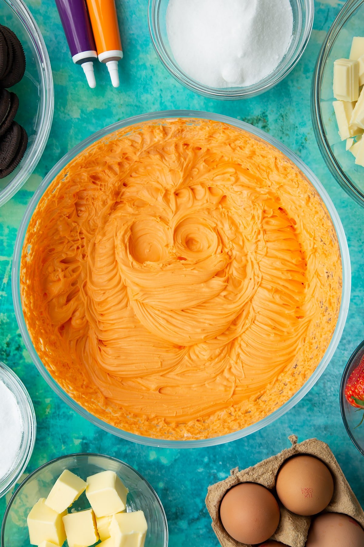 Cream, cream cheese and sugar with orange extract and orange food dye whipped together in a bowl. Ingredients to make Halloween cheesecake surround the bowl.