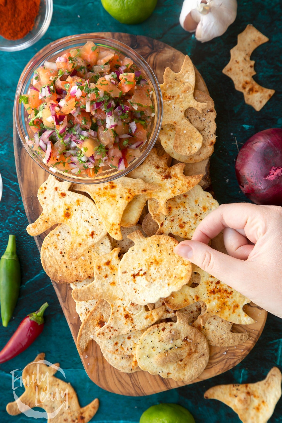 Halloween tortillas on a board with a small bowl of tomato salsa. A hand holds a pumpkin shaped chip.